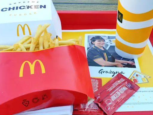Duże frytki McDonald's w czerwonym opakowaniu, obok pudełka z burgerem i kubka z napojem.