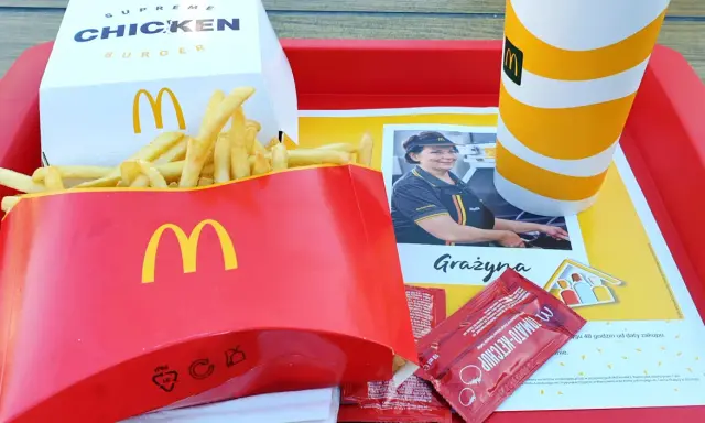 Duże frytki McDonald's w czerwonym opakowaniu, obok pudełka z burgerem i kubka z napojem.